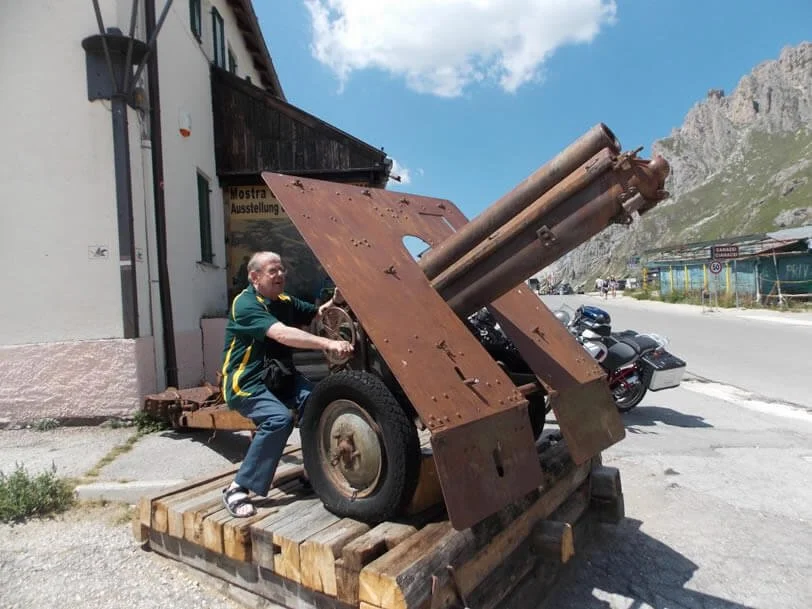 A man is sitting on an old, rusty, large artillery cannon mounted on a wooden platform outdoors with mountains and a blue sky in the background.