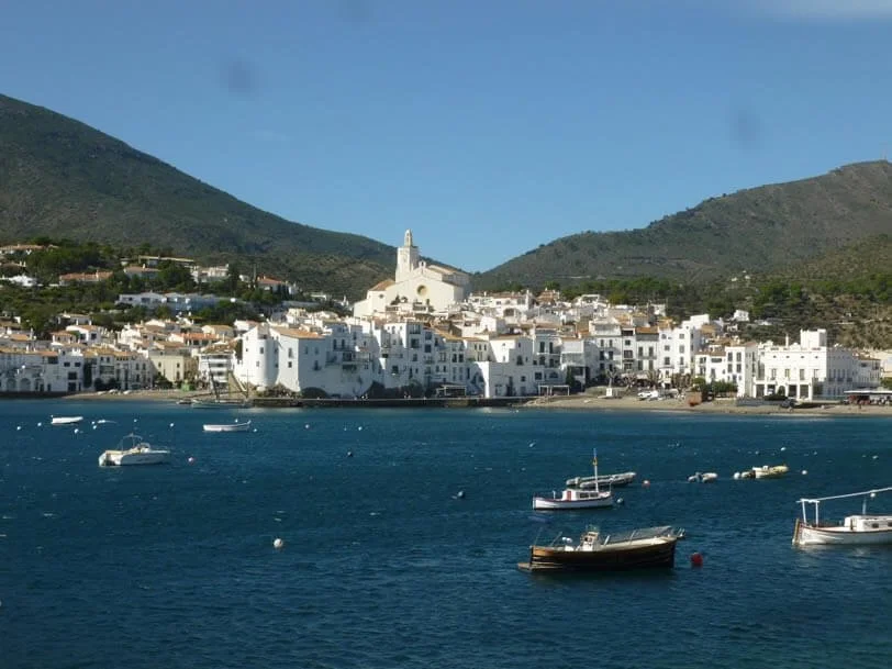 A coastal town with white buildings on a hillside, a church with a tall steeple, and boats in the water.