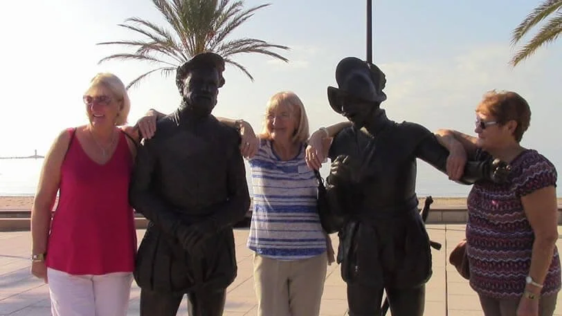 Four women are standing together with two street performers dressed as statues, five palm trees behind, and a beach and ocean in the background during daylight.