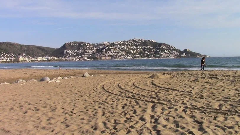 Person walking on a sandy beach with tire tracks, ocean waves, and hills with white buildings in the background.