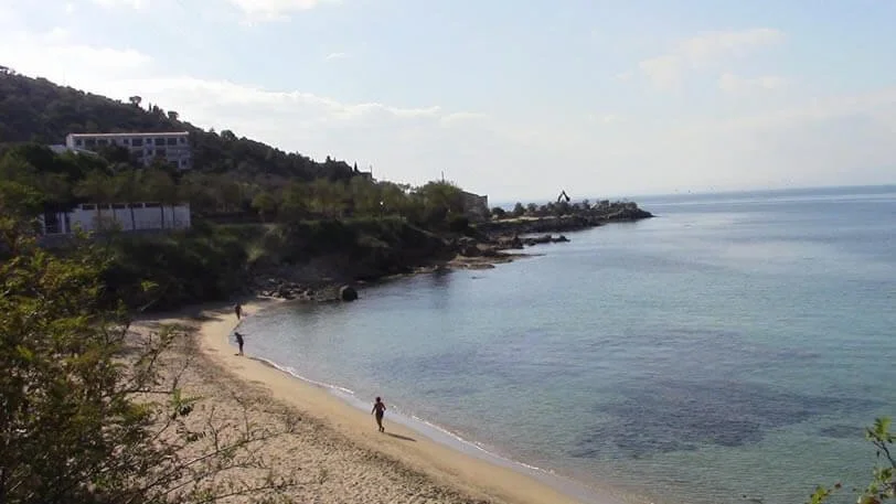 Beach scene with three people walking along the shoreline, rocky coastline, greenery, building on the hill, calm water, and a cloudy sky.