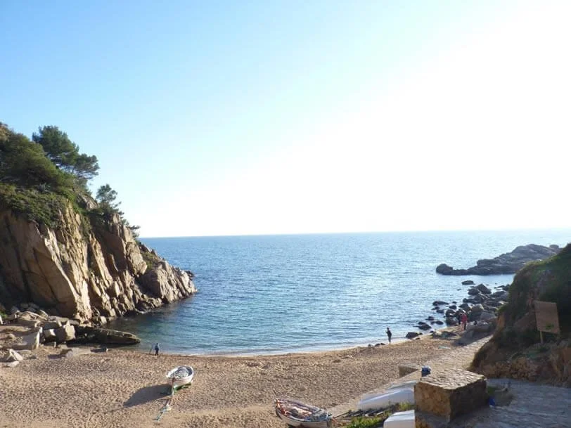 A beach scene with sandy shore, rocky cliffs on both sides, and calm ocean water, with a few people walking.