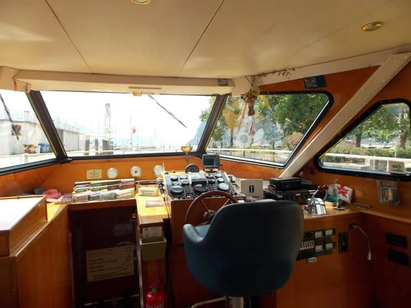 Interior of a boat's bridge or helm, with navigational and communication equipment, a steering wheel, and a black chair, looking out through large front and side windows at outdoor scenery.