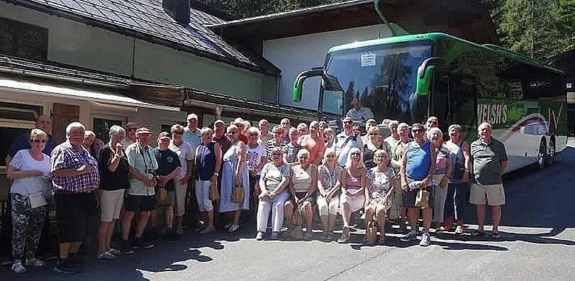 Group of elderly people standing and sitting in front of a large green and white tour bus outdoors.