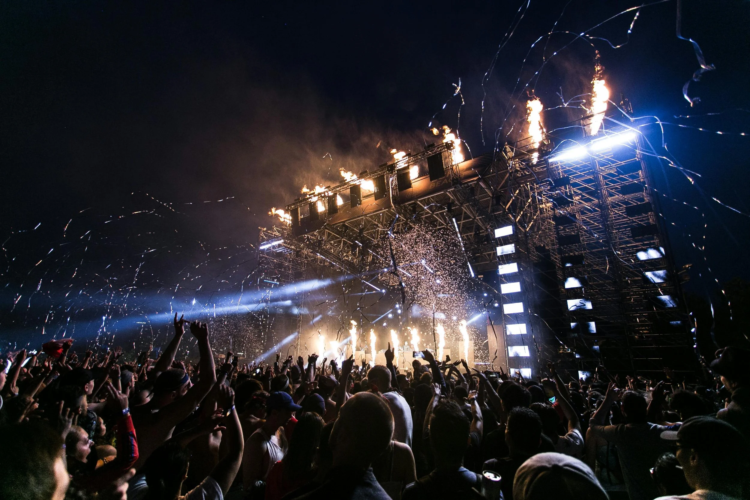Crowd of people at a nighttime outdoor concert, with fireworks, confetti, and stage lighting illuminating the scene.