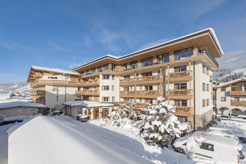 A multi-story hotel in a snowy mountain landscape with wooden balconies and snow-covered cars parked in front.