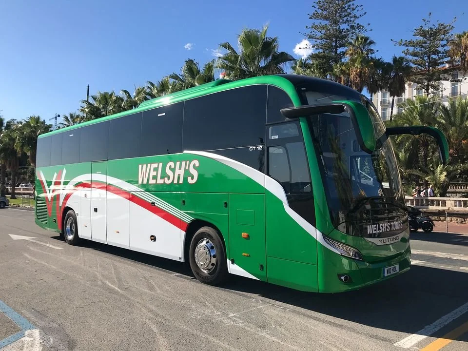 Green and white tour bus with 'Welsh's' branding parked on a street with palm trees in the background.