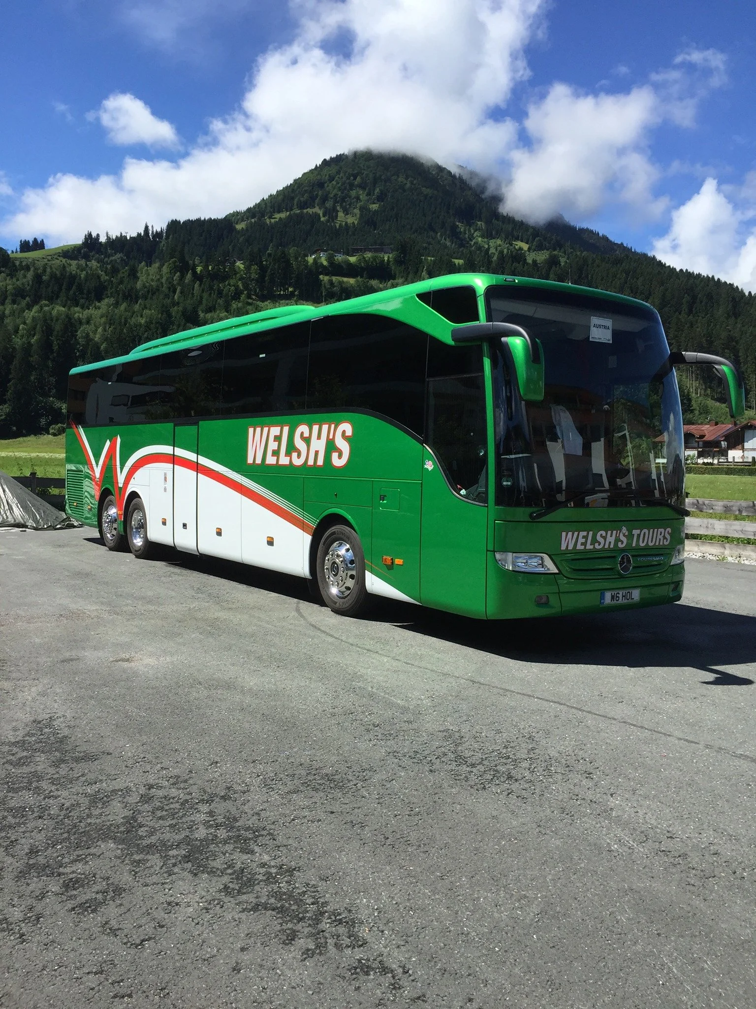 Green tour bus with 'Welsh's Tours' written on the side parked on a paved road with mountains and blue sky in the background.