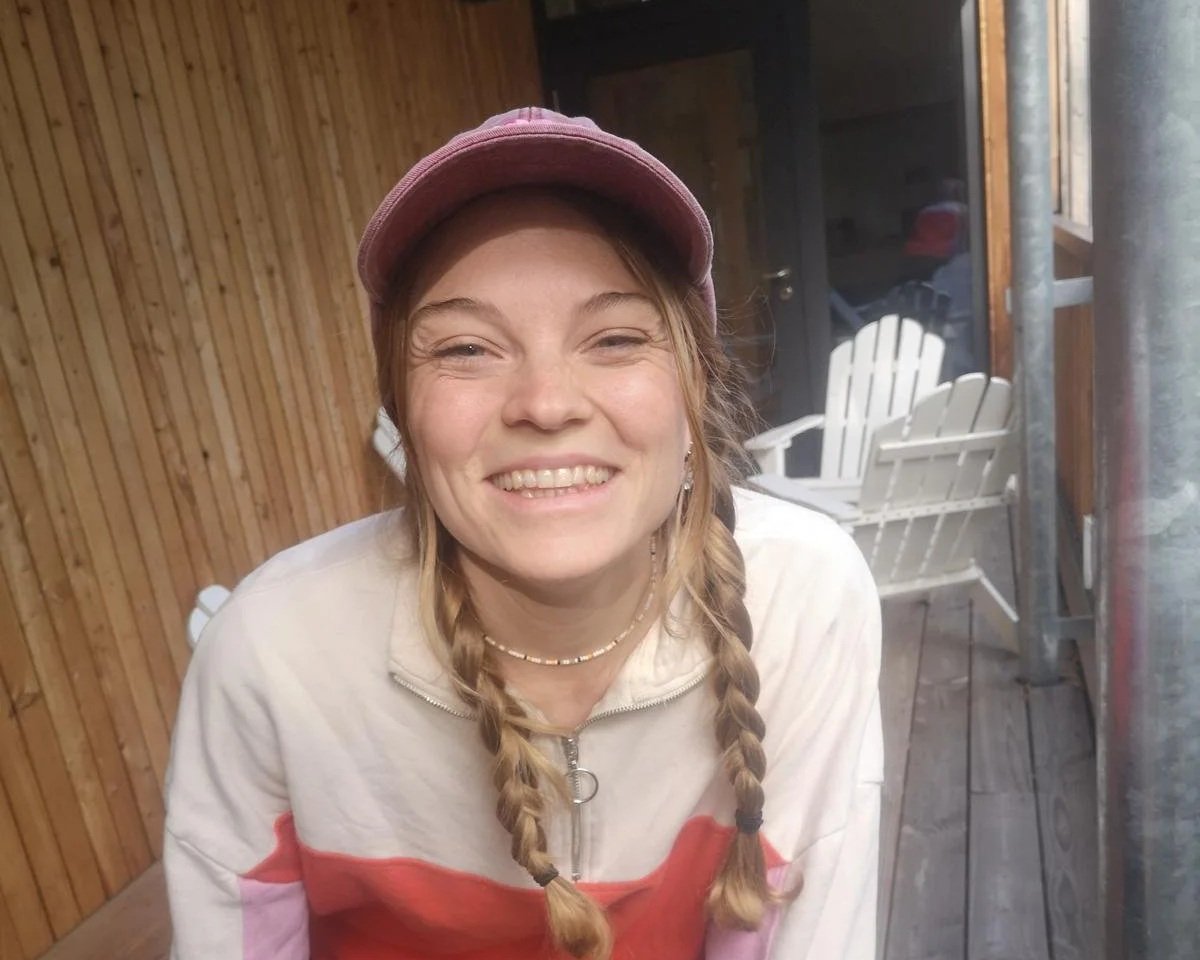Junge Frau im Freizeitoutfit mit Zopf, lila Mütze, braunen Haaren, freundlichem Lächeln, Sitzposition, auf Holzterrasse vor Holzwand, im Hintergrund weiße Gartenstühle, Fenster und Tür.