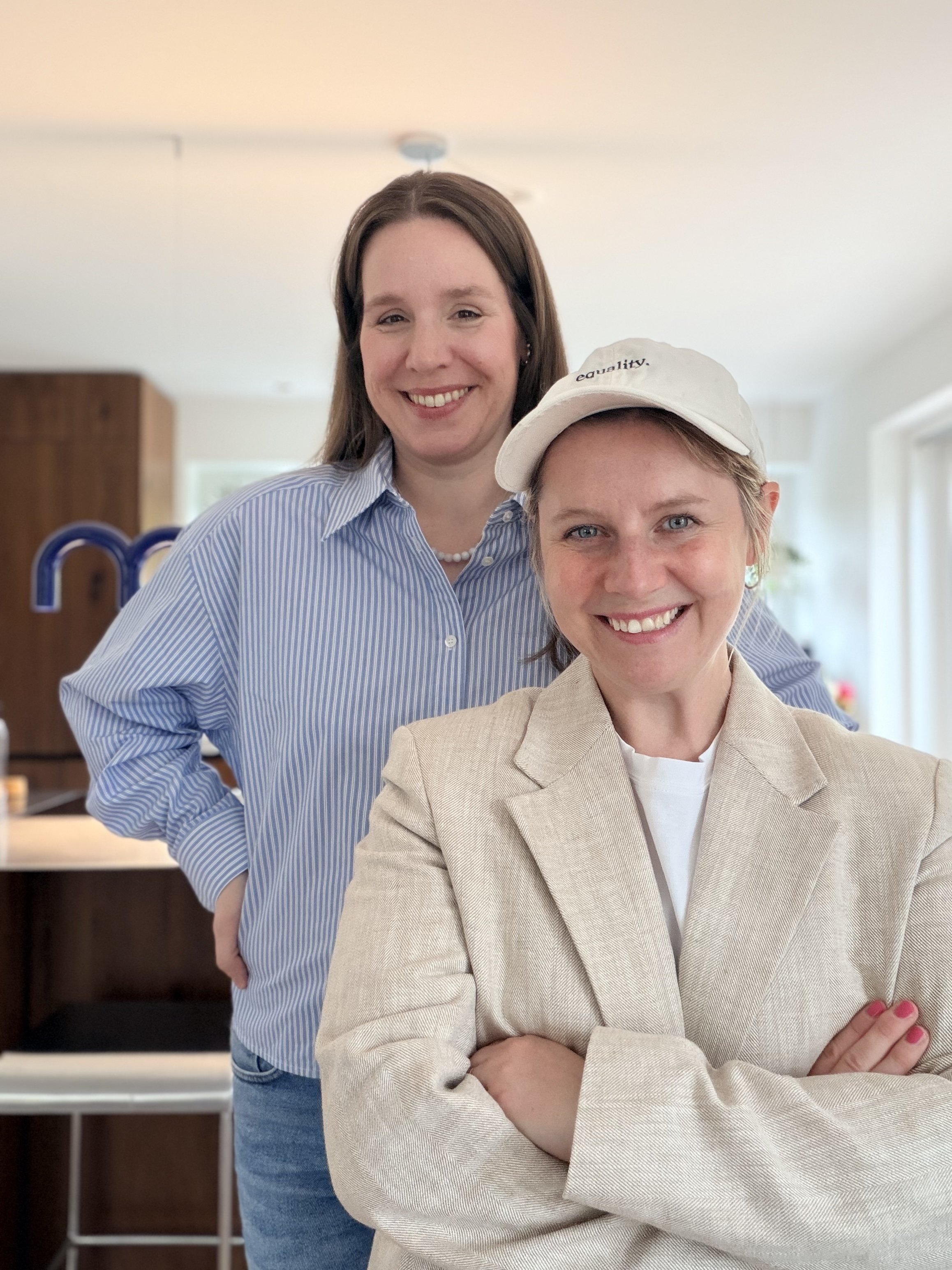 Zwei Frauen, lachend, in einem hellen Raum, eine trägt eine Kappe mit dem Wort ‚equality‘, die andere trägt eine blaue gestreifte Bluse.