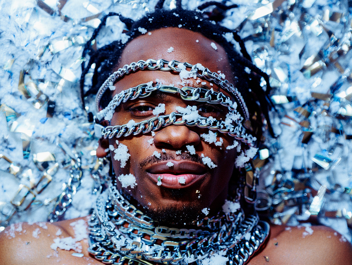 A person lying down covered in silver metallic tinsel, wearing multiple silver chains around the neck and forehead, with snowflakes on the face and chains, and a small piercing on the lip.