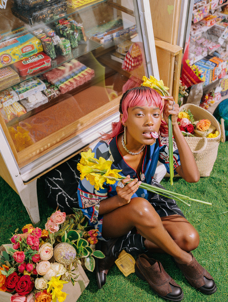 A person with pink hair sitting on artificial grass floor inside a store, surrounded by flowers and flowers arrangements, holding yellow daffodils, with candy and snacks visible in a refrigerated display case behind them.