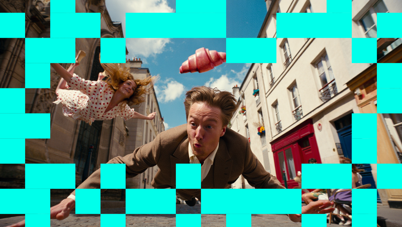A young man in a brown suit with light hair is lying on the street, looking at the camera with a surprised expression. A young woman in a polka dot dress is mid-air, falling or floating behind him. The scene takes place on a city street with buildings, balconies, and a blue sky with clouds.