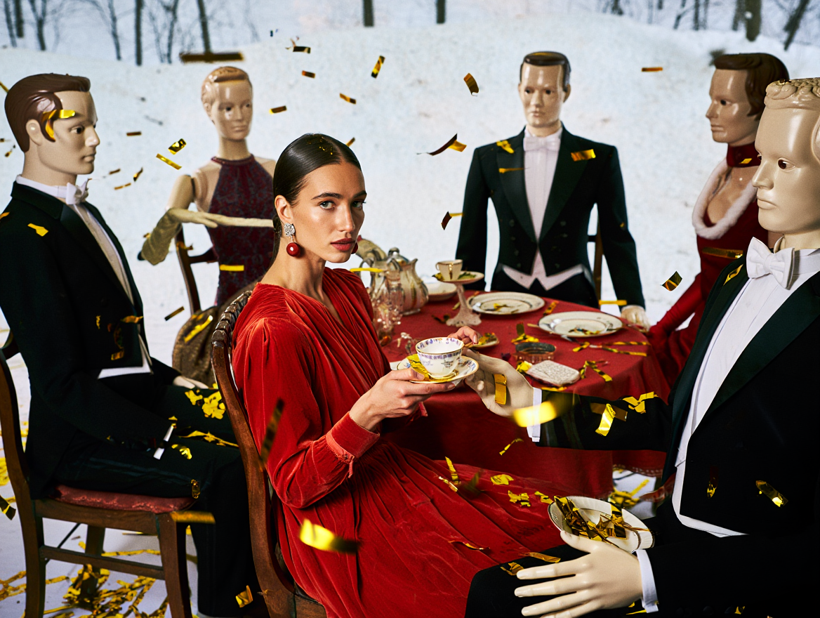 A woman in a red dress at a formal dinner table, surrounded by male and female mannequins dressed in tuxedos and evening gowns, with confetti falling around them.