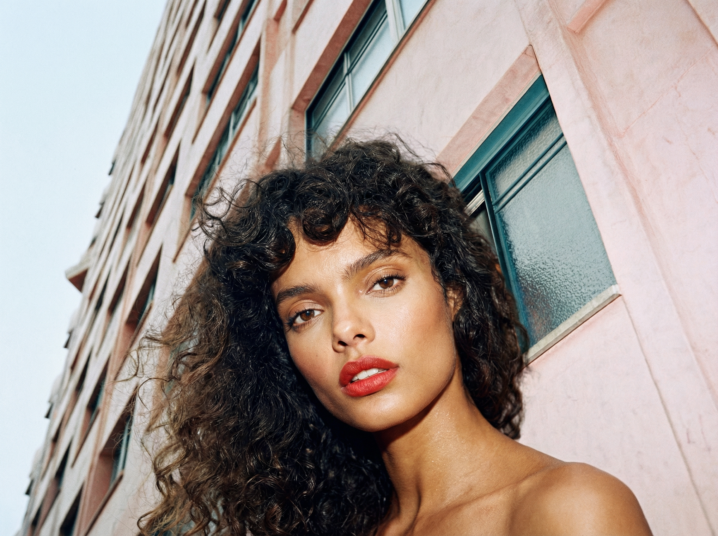 Close-up of a woman with curly dark hair and red lipstick standing against a pink building with multiple windows.