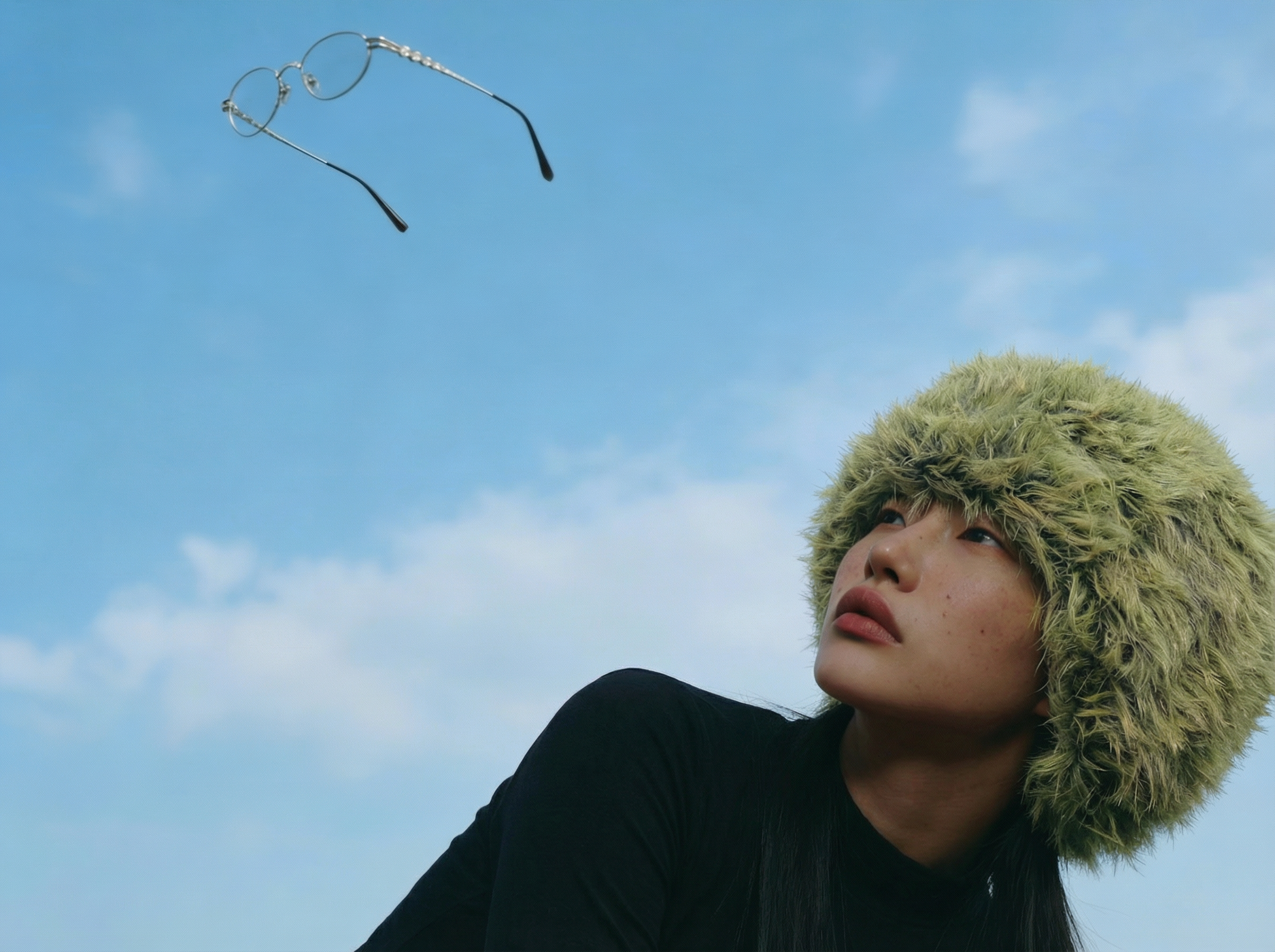 A woman wearing a fuzzy, green hat looking up at a pair of glasses flying through the sky against a backdrop of blue sky and clouds.