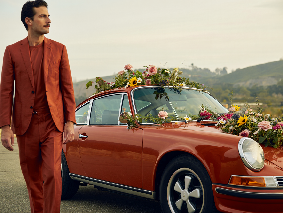 A man in a red suit standing next to a vintage red car decorated with flowers on the roof and hood, set against a scenic outdoor backdrop.