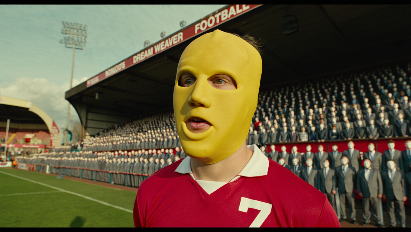 A person wearing a yellow mask and red sports jersey with the number 7 on it, standing on a sports field with a crowd of mannequins dressed in suits behind them.