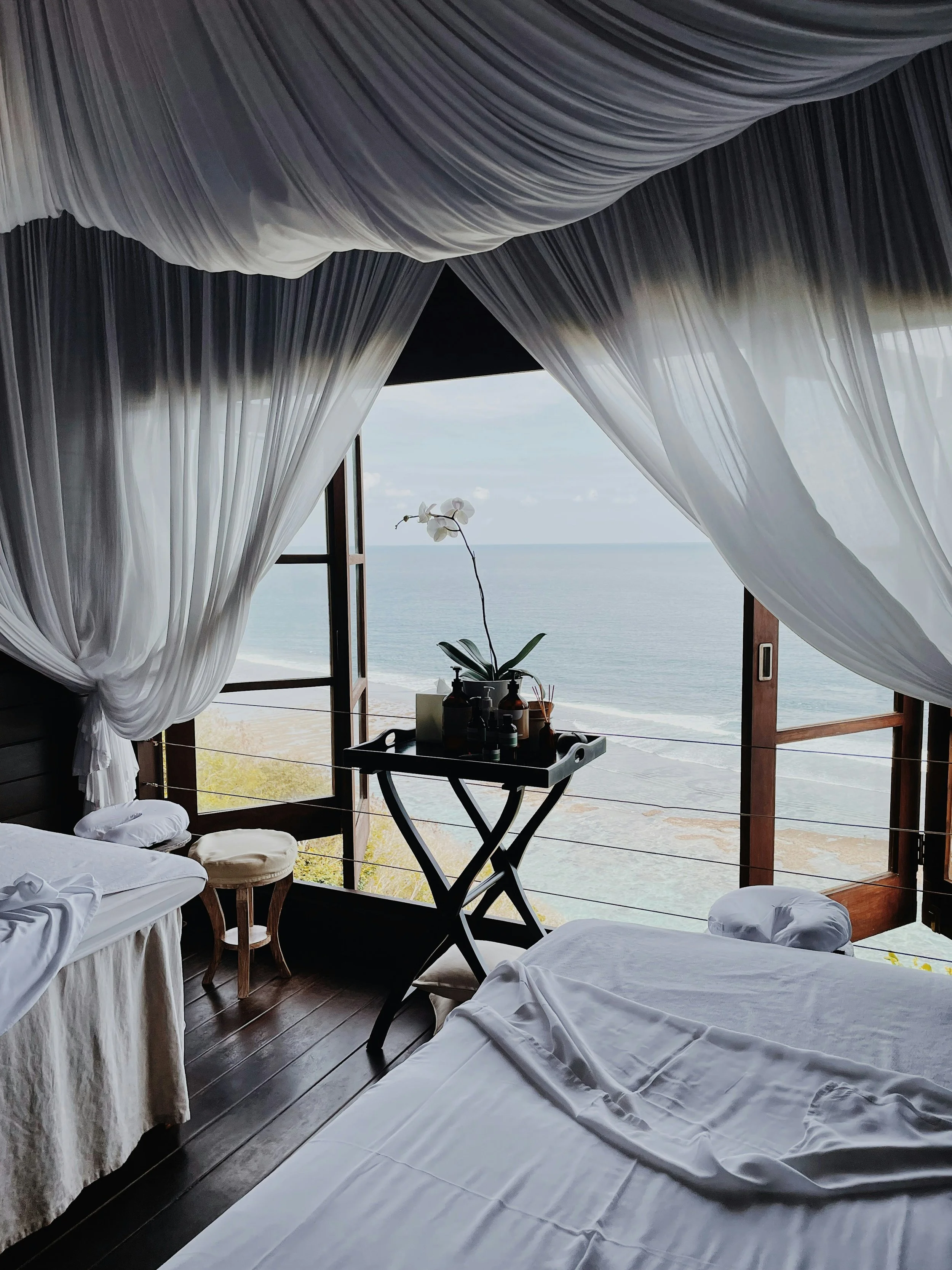 Cozy bedroom with white bedsheets, overlooking the ocean through open wooden window shutters, with sheer white curtains, and a small table holding a potted orchid and toiletries.