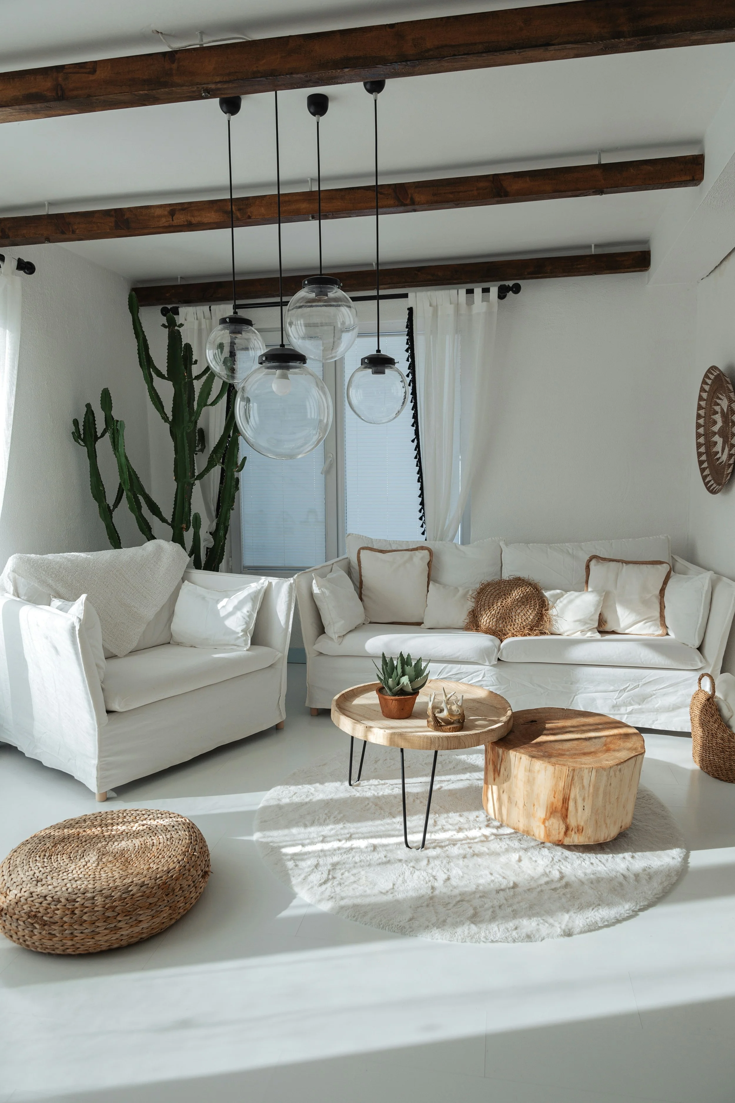 Bright living room with white sofas, a large cactus, hanging pendant lights, a wooden coffee table, a woven pouf, and a round wall art, with sunlight streaming through the window.