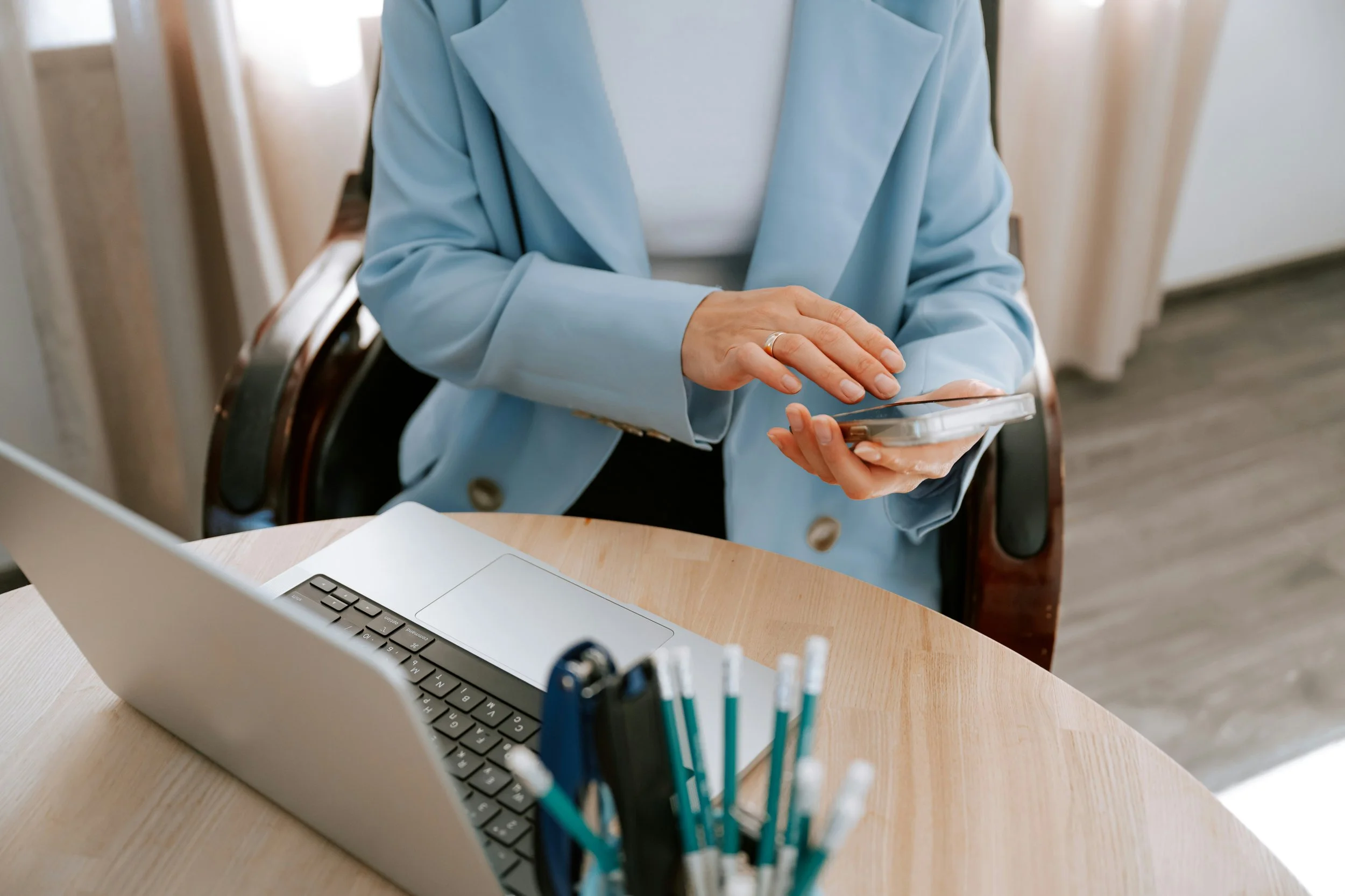 A person in a light blue blazer is sitting at a desk, using a smartphone. On the desk, there is a silver laptop with a black keyboard and a pen holder with several pens.