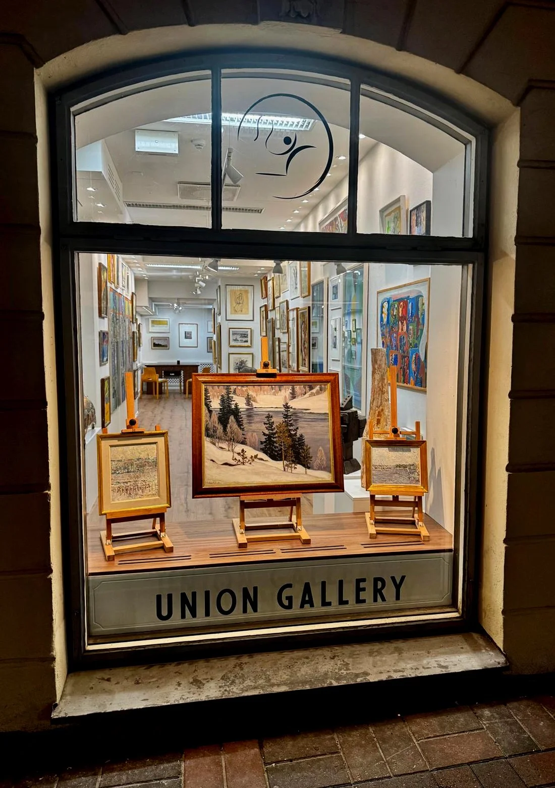 Näky ikkunoista, jossa on taidegalleria, jossa on useita maalauksia ja tauluja. Edessä kolme pienempää maalausta ja keskellä suurempi maalaus, kaikkia kehystyksissä. Gallerian nimi on Union Gallery.