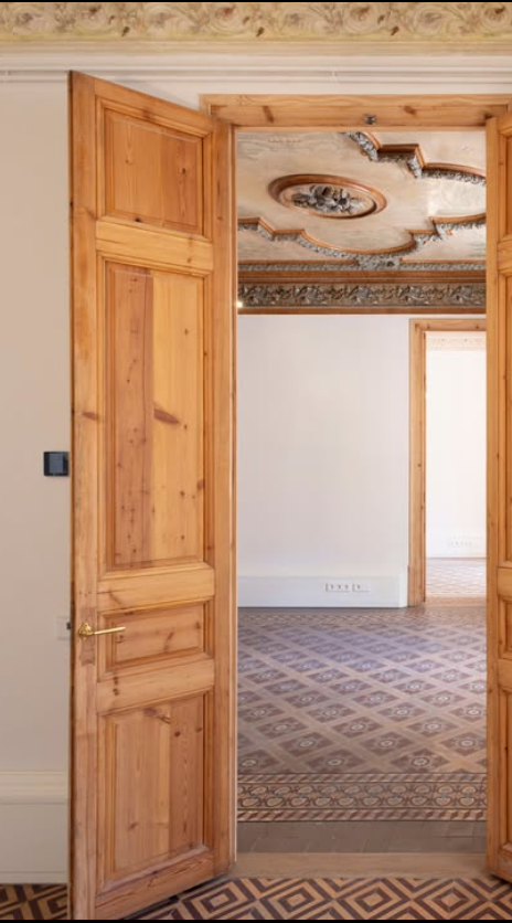 Interior view through wooden double doors into a room with ornate ceiling design and patterned floor tiles.