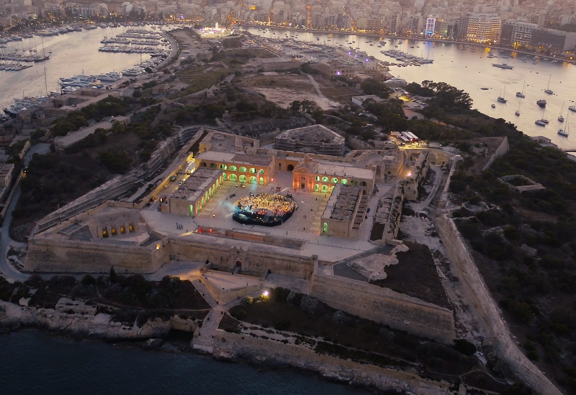 Aerial view of a historic fortress with a stage set up inside, illuminated with green and yellow lights, overlooking a harbor with boats and a city skyline at dusk.