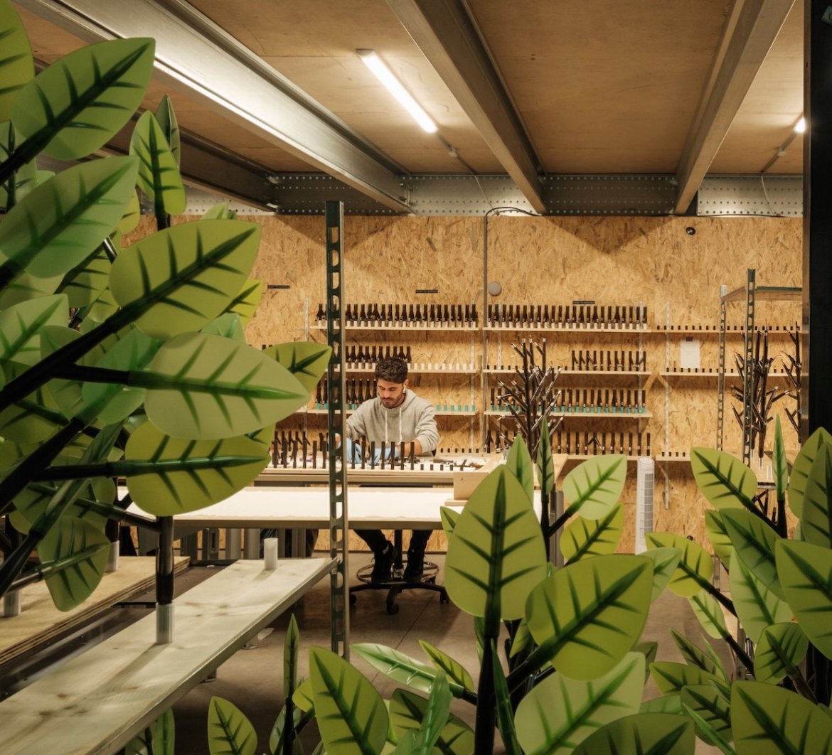 An architect man focused, creative, smart, dedicated, artwork, production, technical, detailed, high end, working at a crafting table in a workshop with shelves of tools and supplies in the background, framed by green leafy plants in the foreground.