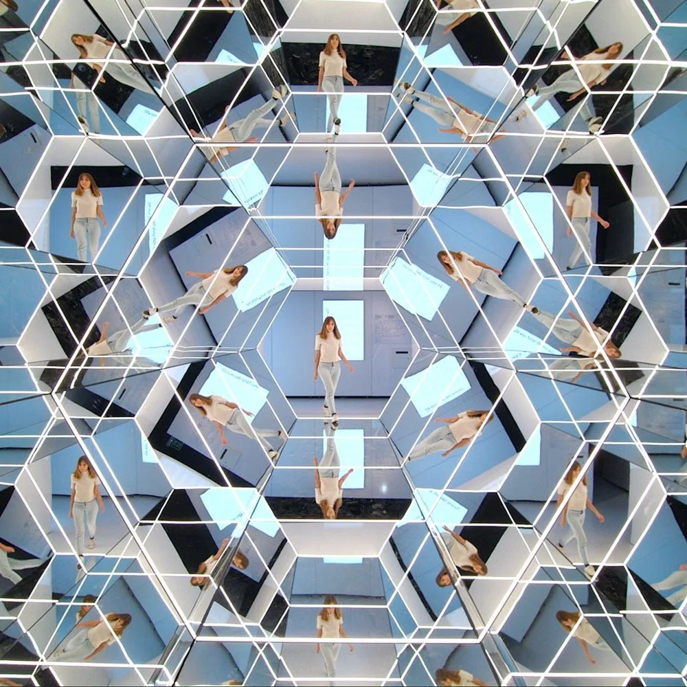 A woman standing in the middle of a room surrounded by mirrors forming a complex geometric pattern reflecting her image repeatedly.