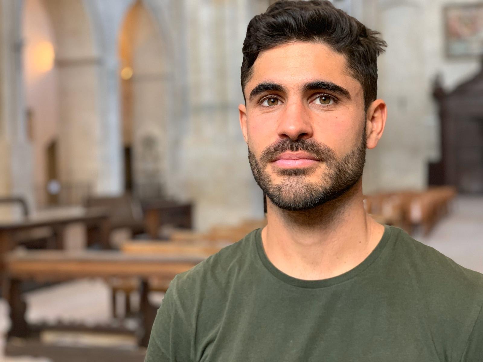 A man with dark hair and a beard, wearing a green t-shirt, standing inside a church with stone columns and wooden pews in the background.