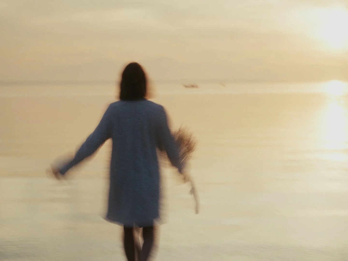A bride walking toward the sea during sunset or sunrise, holding a bouquet of flowers.