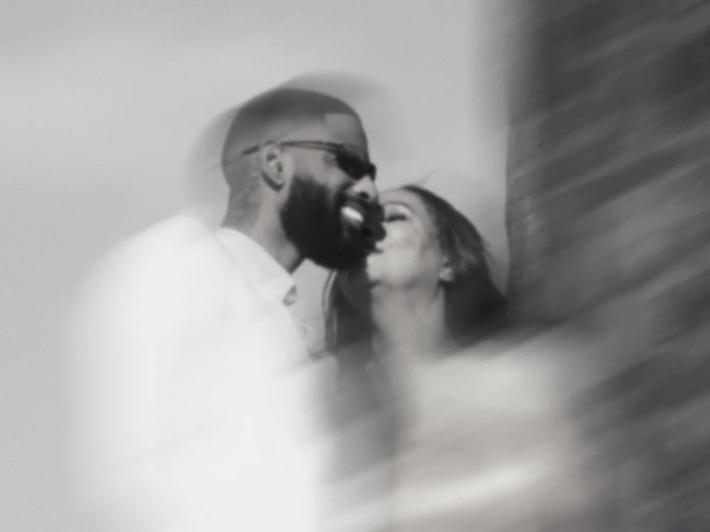 Black and white photo of a man and woman laughing close together, with the man wearing sunglasses and having a beard.