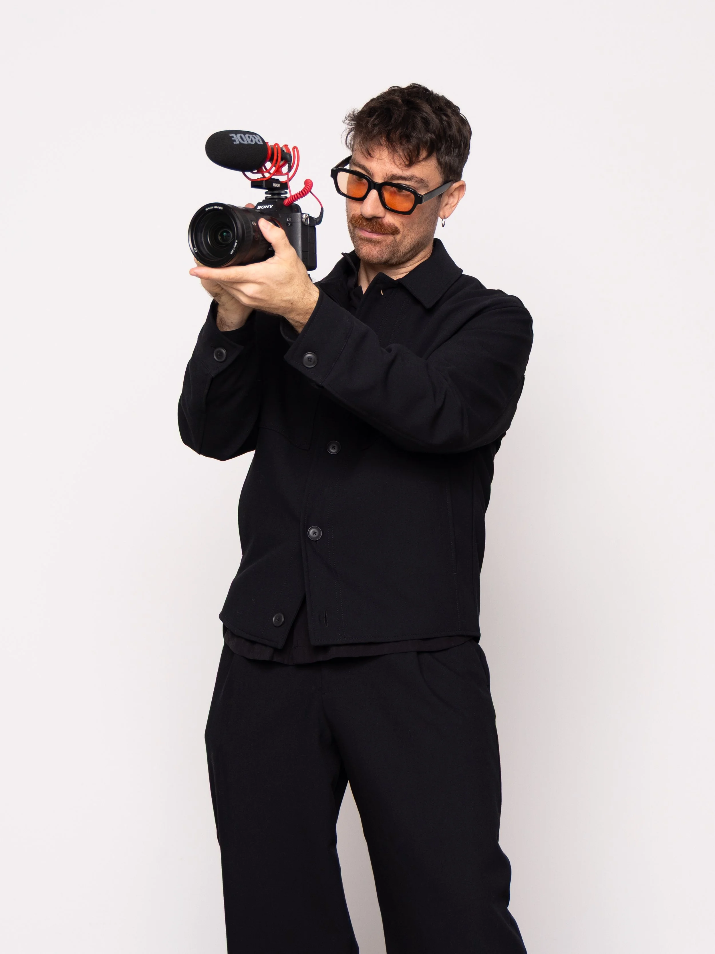 A man wearing black clothing, glasses, and earrings holding a professional video camera with a microphone attached.