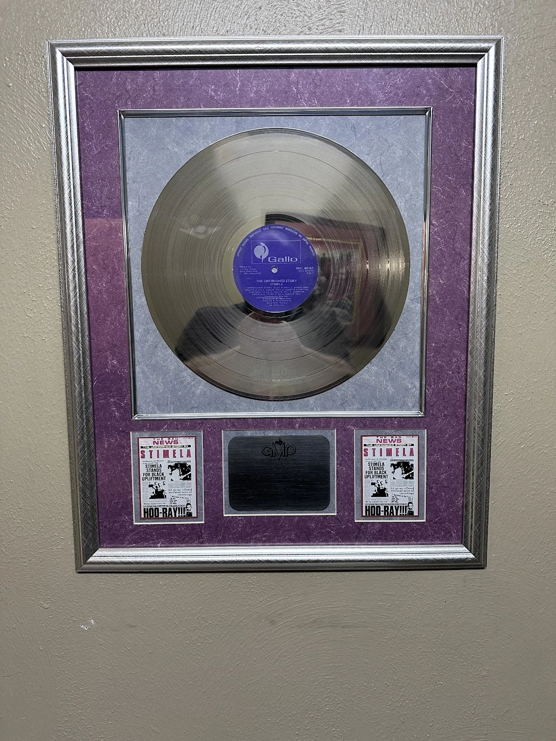Framed platinum record award with two small newspaper clippings at the bottom and a silver plaque in the center, mounted on a beige wall.