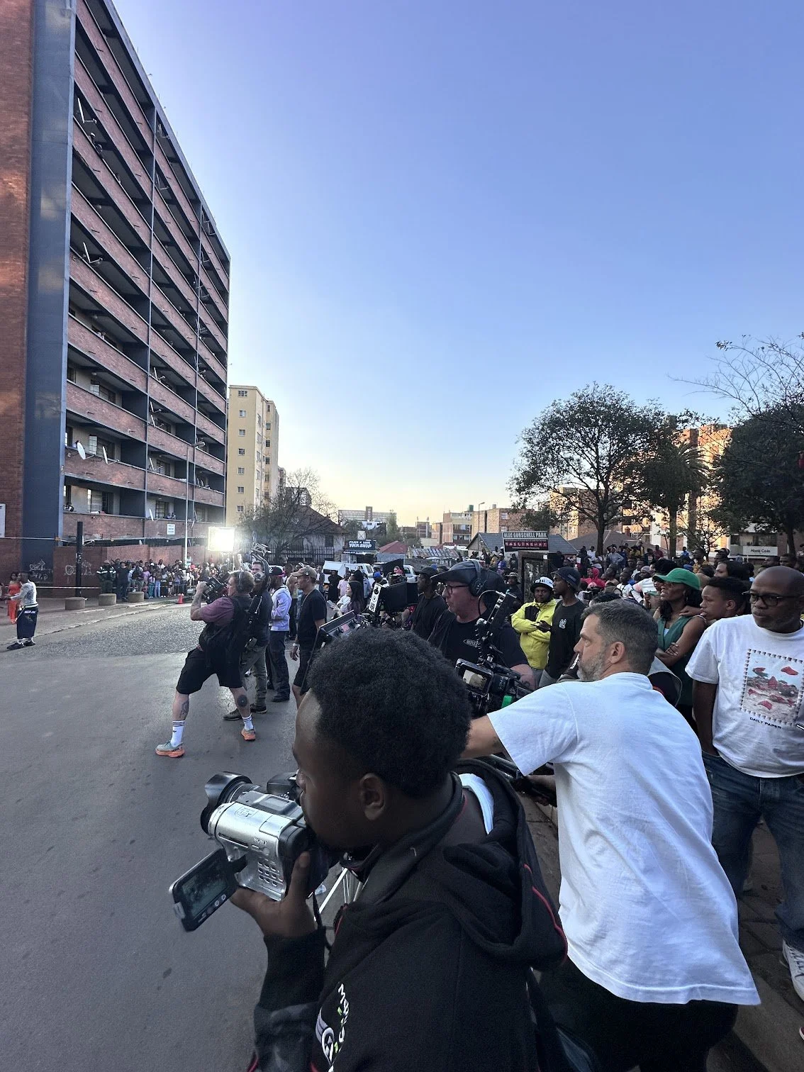 A crowd of people, including news reporters and photographers, gathering outside a building in an urban area with high-rise apartments on the left. Some are recording or taking pictures, while others watch. Trees and additional buildings are visible 