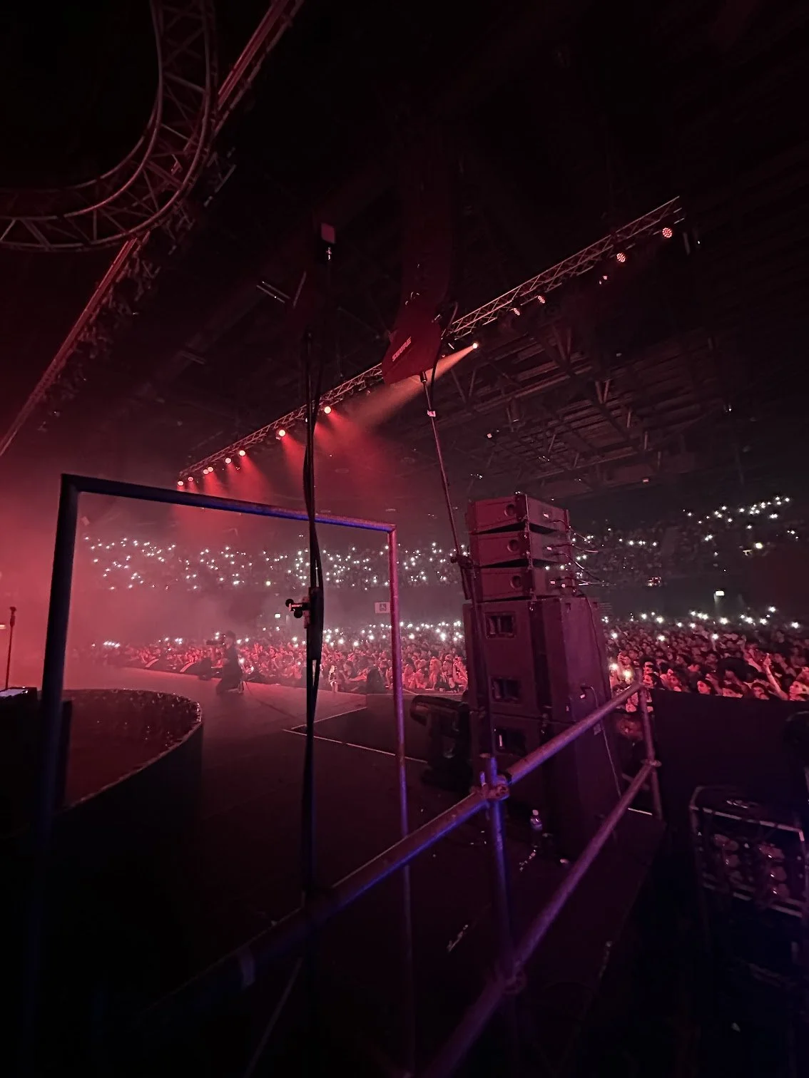 Photo taken from the stage at a concert, showing the audience with many lights and the stage with red lighting.