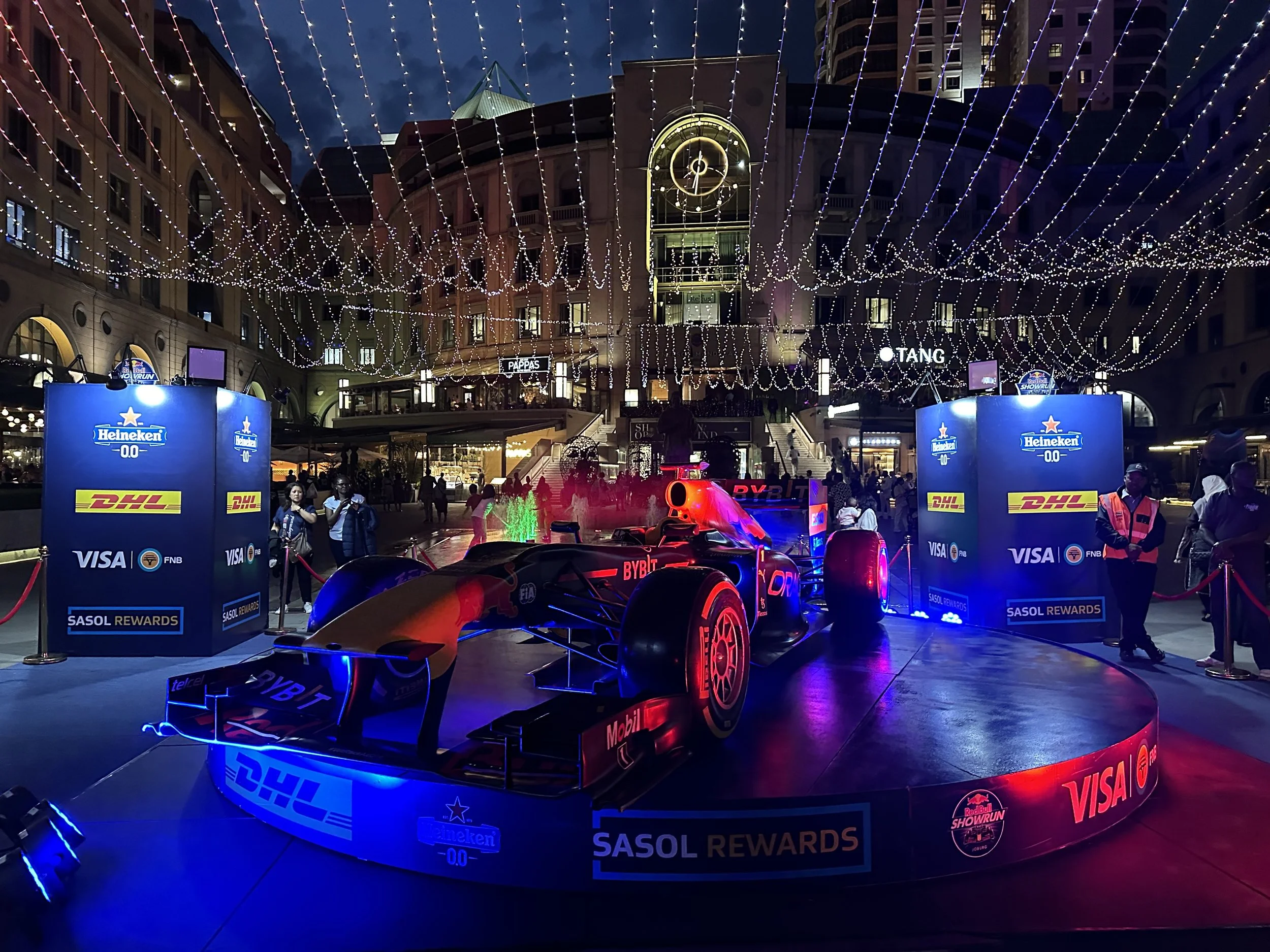 A Formula 1 race car on display in an outdoor shopping center decorated with string lights at night. The car is illuminated with red and blue lighting and is surrounded by sponsor banners, including Heineken, DHL, Visa, and SASOL Rewards.