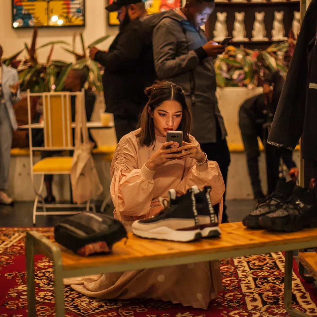 Young woman sitting cross-legged on the floor, looking at her phone, with sneakers on a table in front of her, in a busy indoor setting with several people, plants, and artwork in the background.