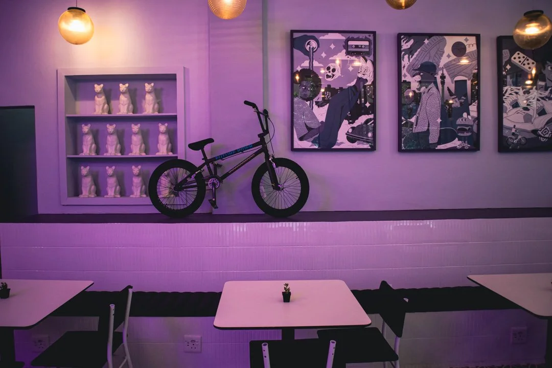 Interior of a cafe with purple lighting, decorated with a bicycle, a shelf with nine cat statues, and three framed artworks on the wall, with tables and chairs in the foreground.