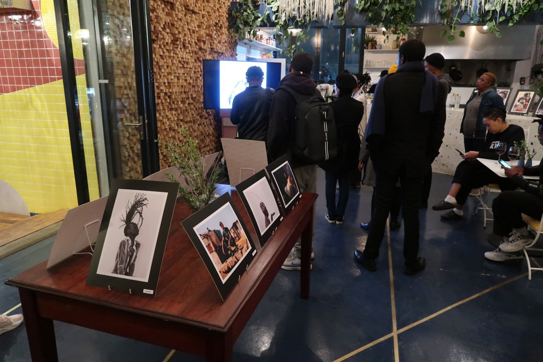 An art exhibition in a modern indoor space with people viewing photographs displayed on a table and on a television screen. The photographs include a black-and-white portrait with a plant headdress, and others with colorful scenes. The space has plan