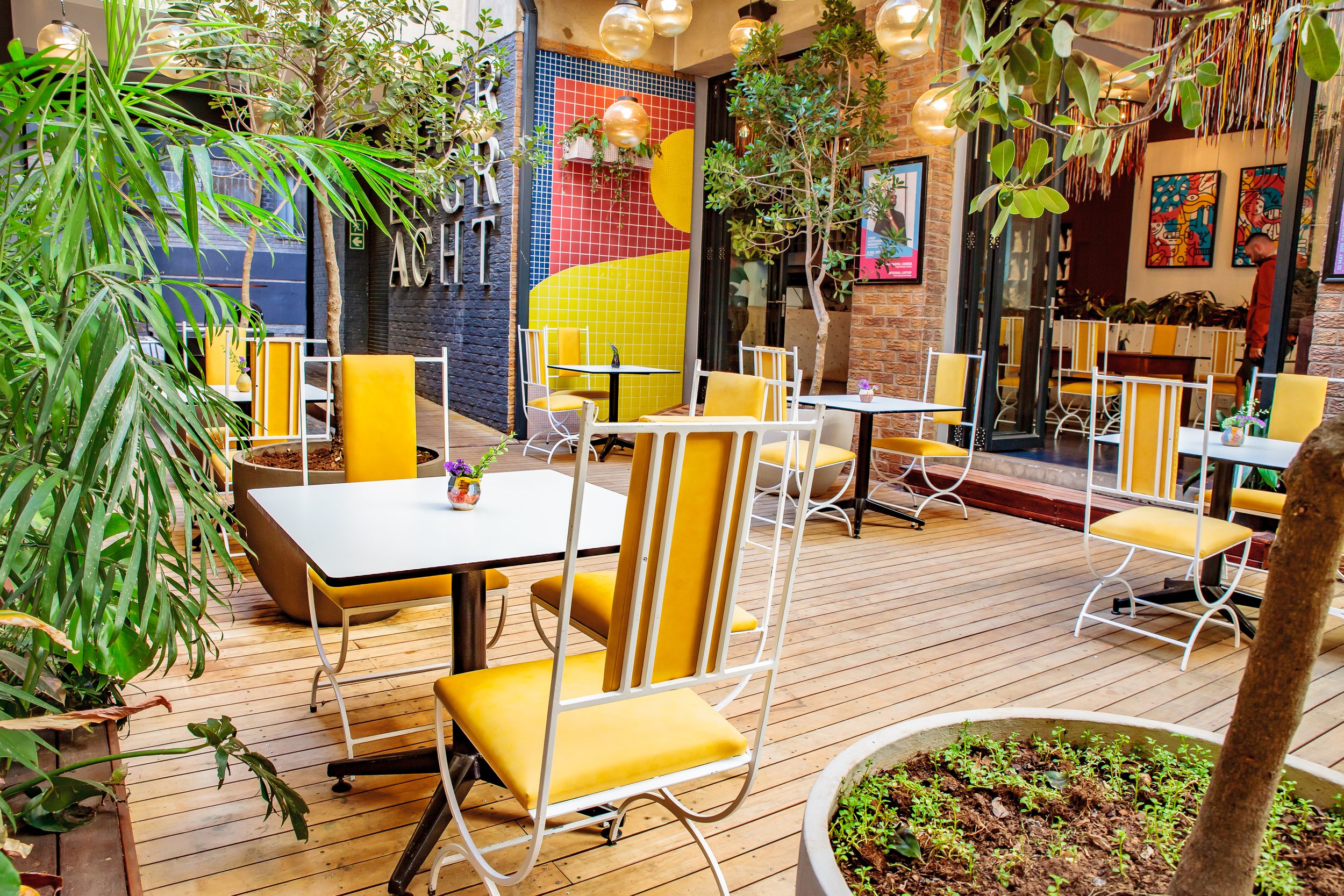 Outdoor seating area of a cafe with yellow chairs, white tables, potted plants, string lights, and colorful wall art.
