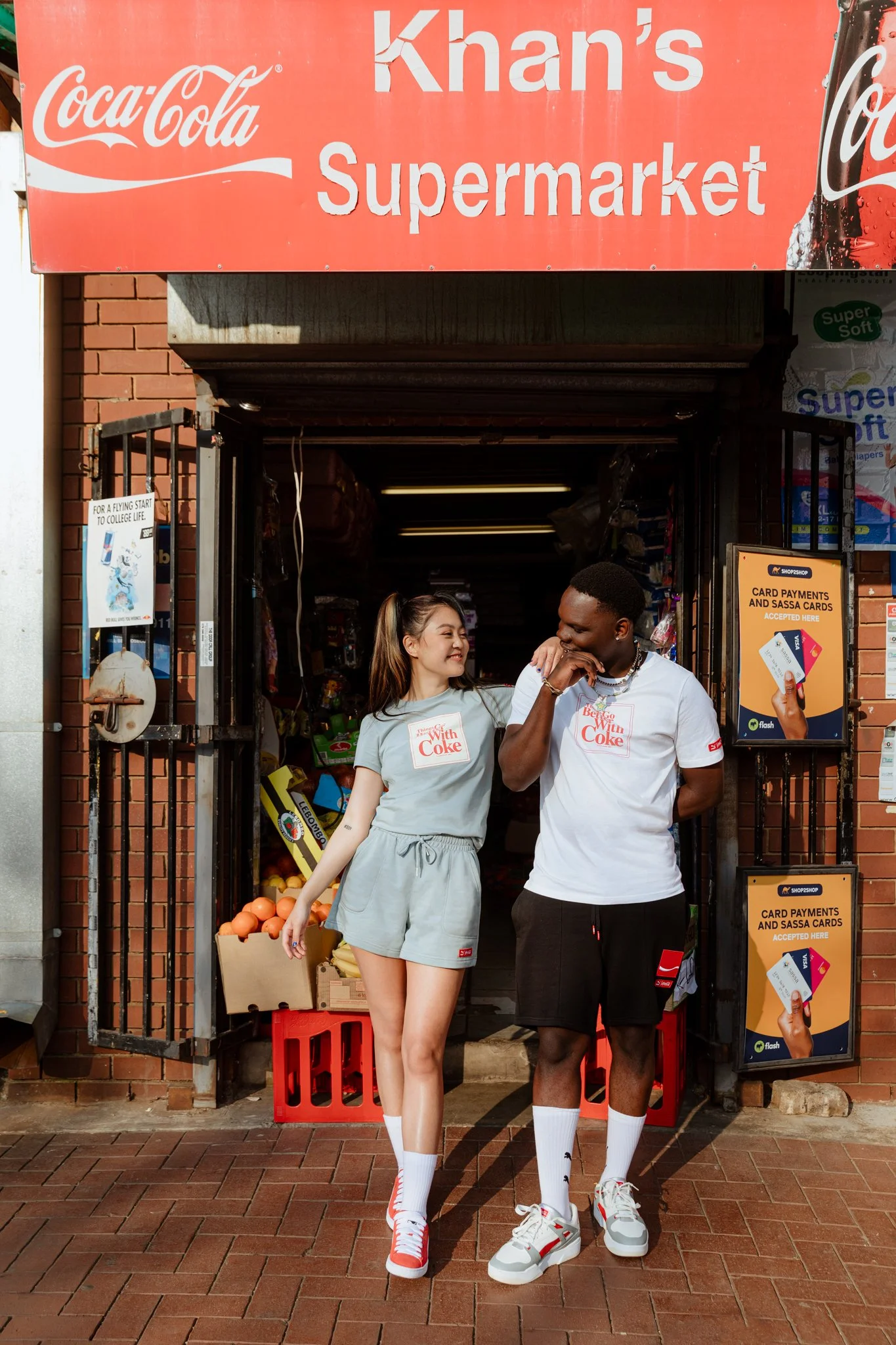 Two friends talking outside Khan's Supermarket, standing in front of the store entrance. The woman is wearing a gray t-shirt and shorts, and the man is in a white t-shirt and black shorts. They are smiling and sharing a moment of laughter. The store 