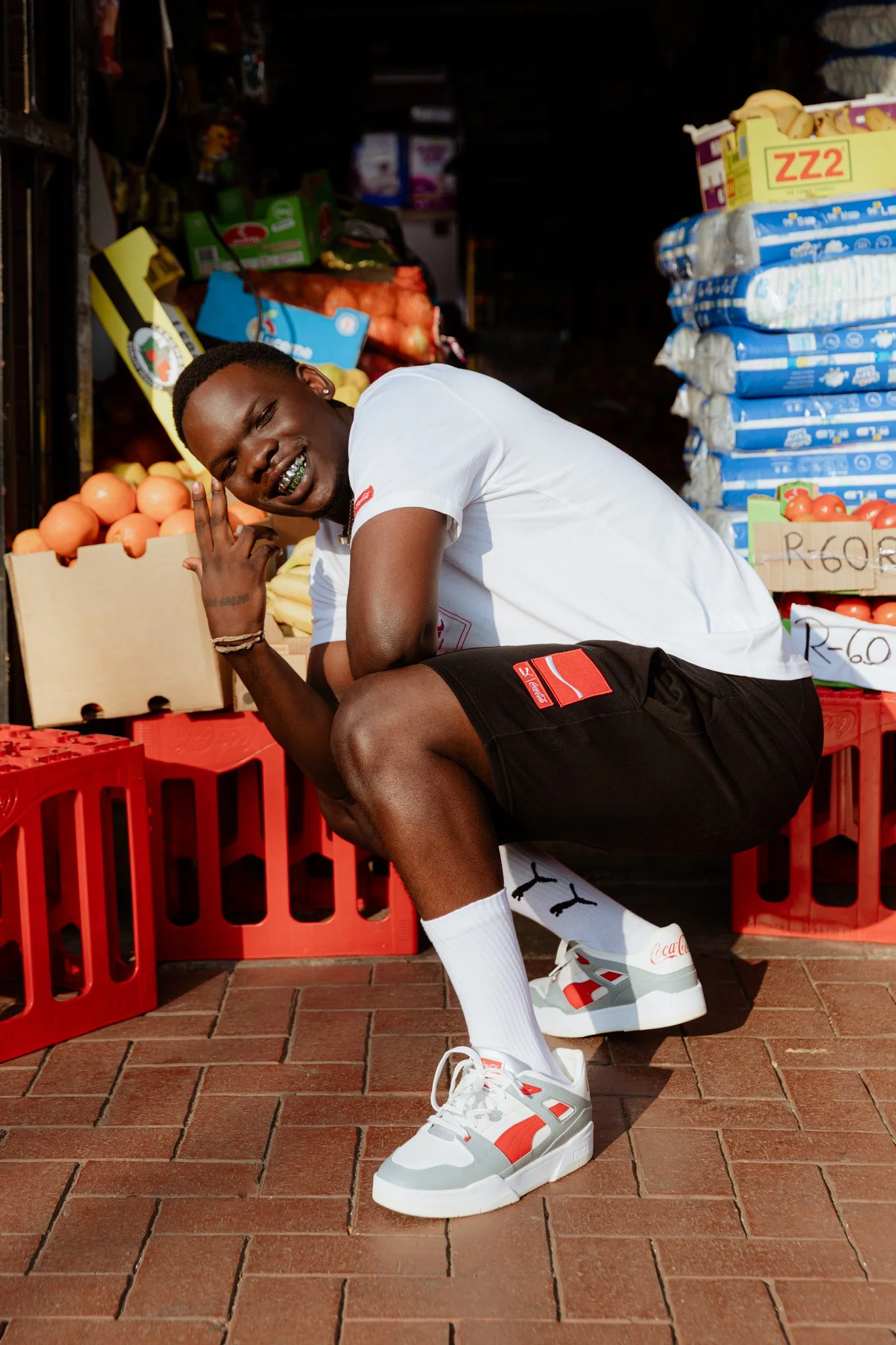 A young man is squatting in front of a fruit and vegetable stand, smiling, wearing a white t-shirt, black shorts, white sneakers with red accents, and white socks, with produce and packaged goods in the background.