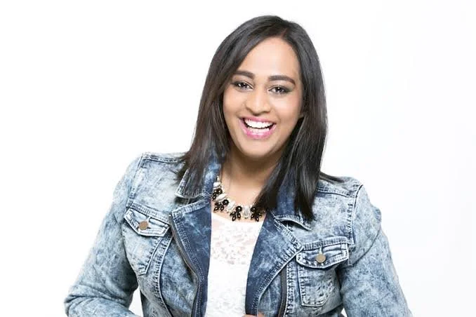 A young woman with medium-length dark hair, smiling, wearing a denim jacket and a black necklace against a white background.
