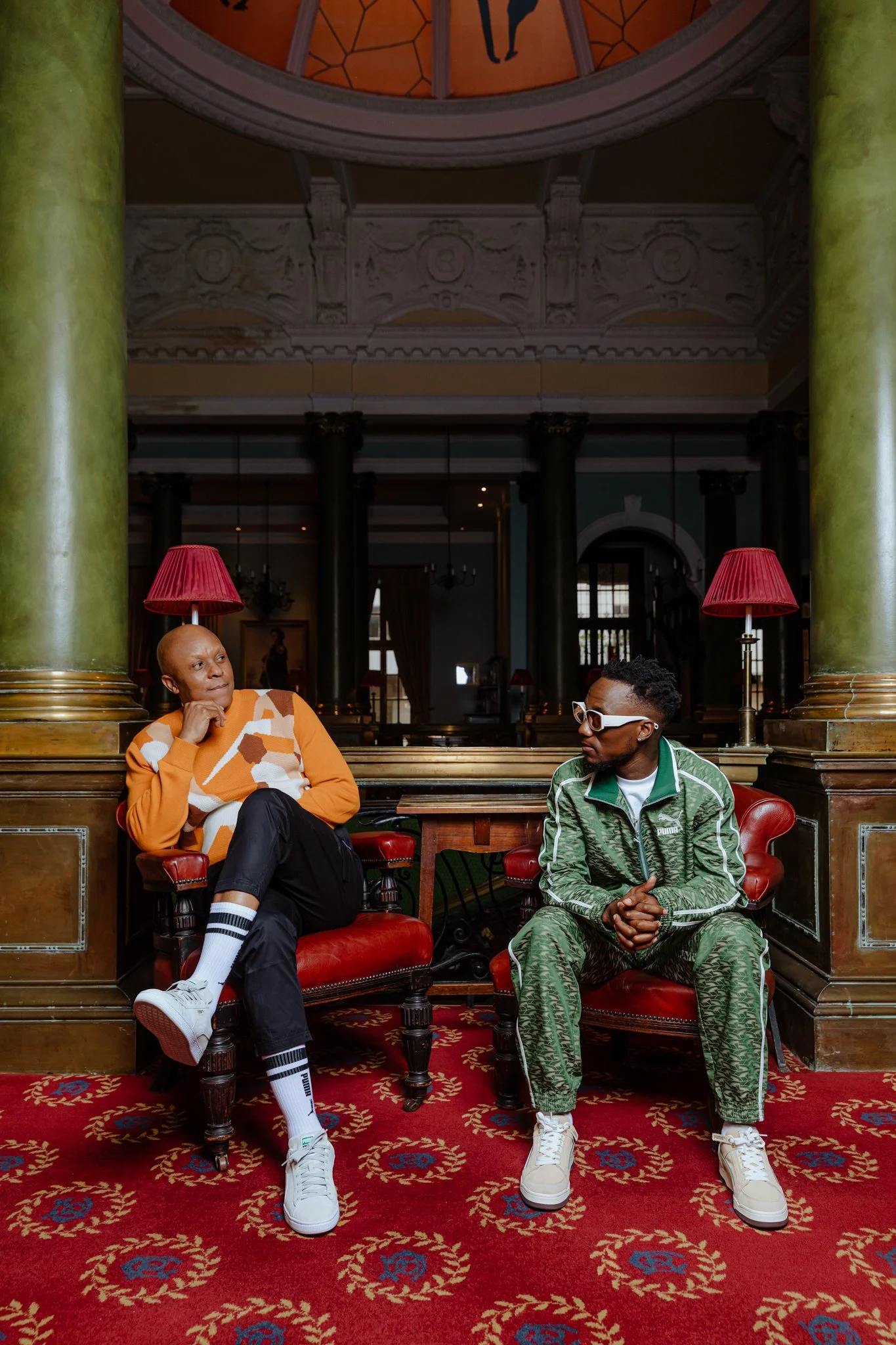 Two men are sitting and conversing in an elegant, vintage-style room with large green columns, red lamps, and a patterned red carpet. One man is wearing a patterned sweater, black pants, and white sneakers, while the other is dressed in a green track