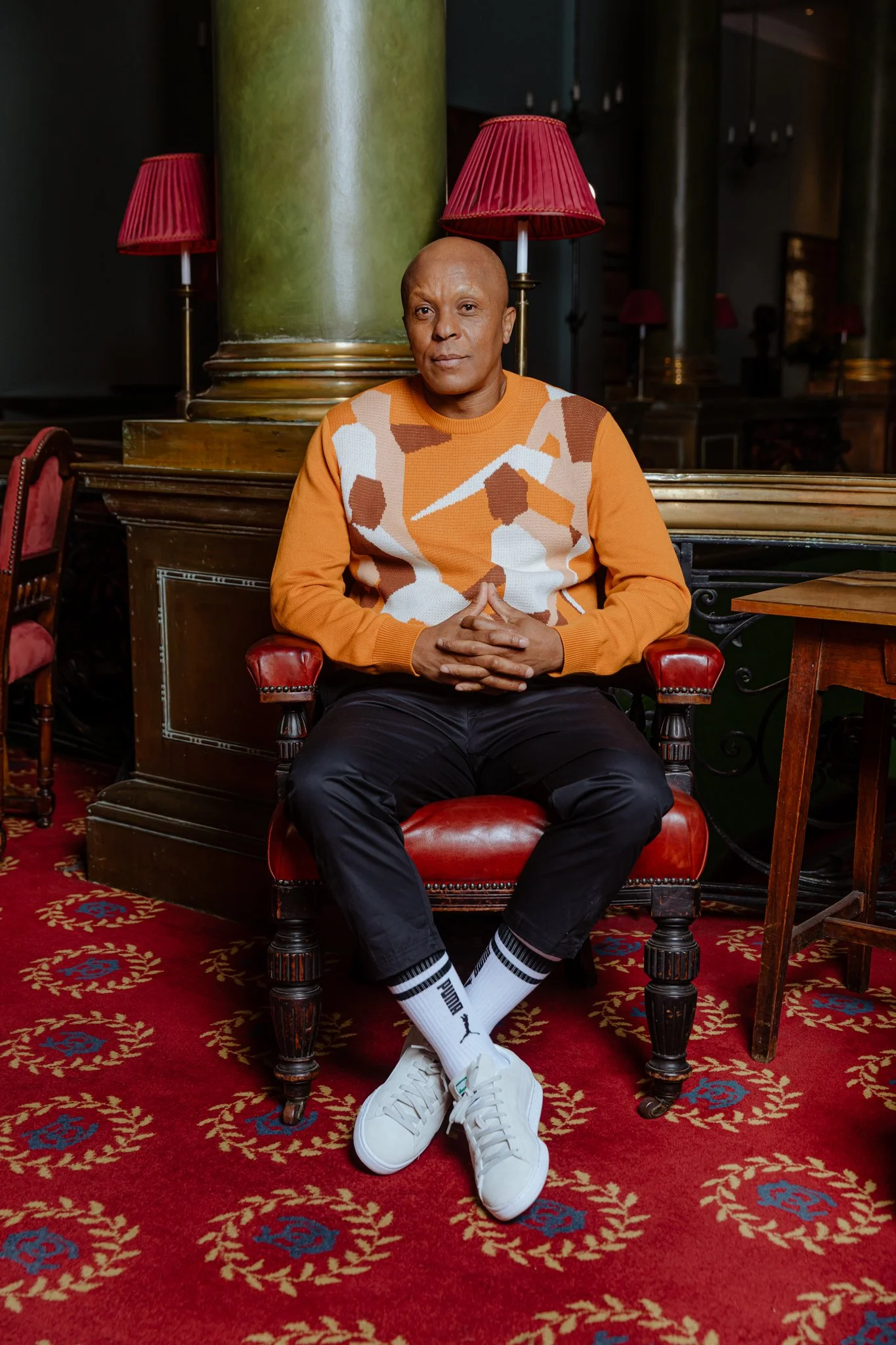 A man sitting on a red leather chair in a luxurious room with wooden furniture, green columns, and red lamps in the background, wearing an orange and white patterned sweater, black pants, white Puma sneakers, and striped Puma socks.