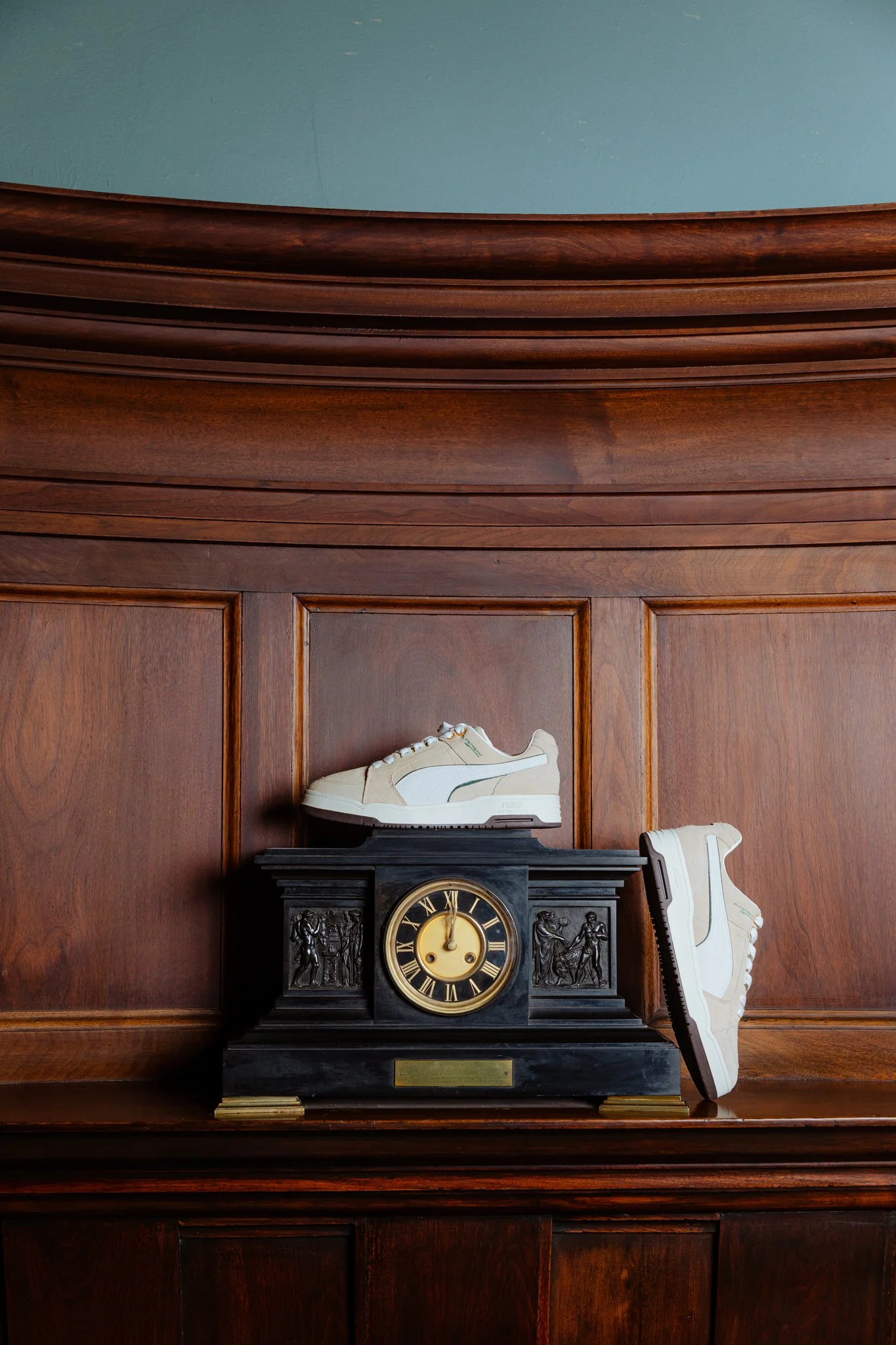 An antique black clock with gold Roman numerals and decorative reliefs on either side sits on a wooden surface. Two beige and white sneakers are placed on top of and beside the clock.