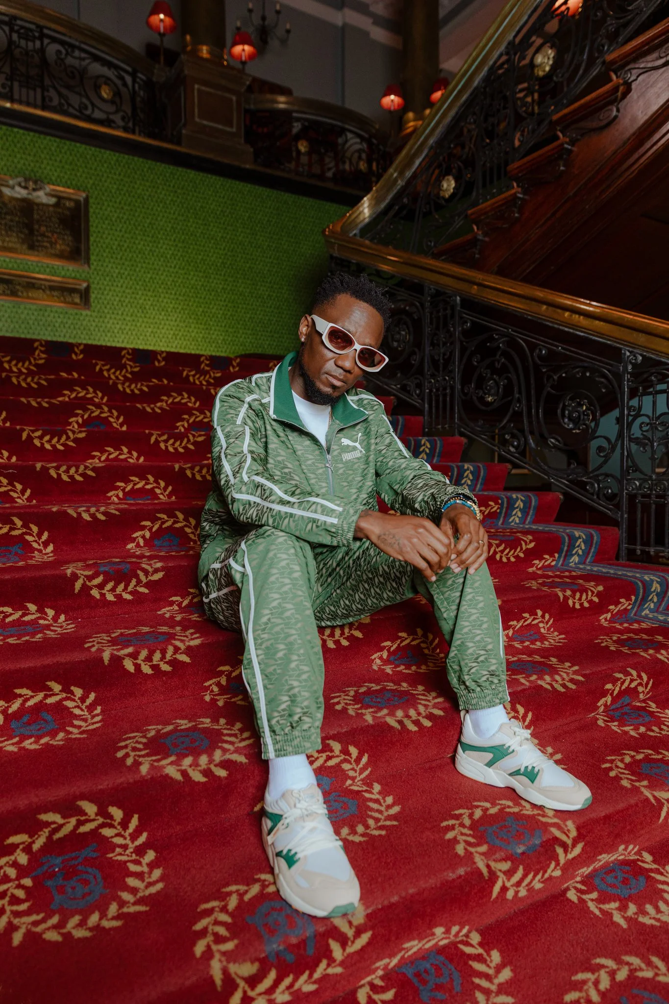 A man dressed in a green Puma tracksuit with white and green sneakers, sitting on a decorative red carpeted staircase inside a stylish, vintage interior with ornate black railings and red lamps.
