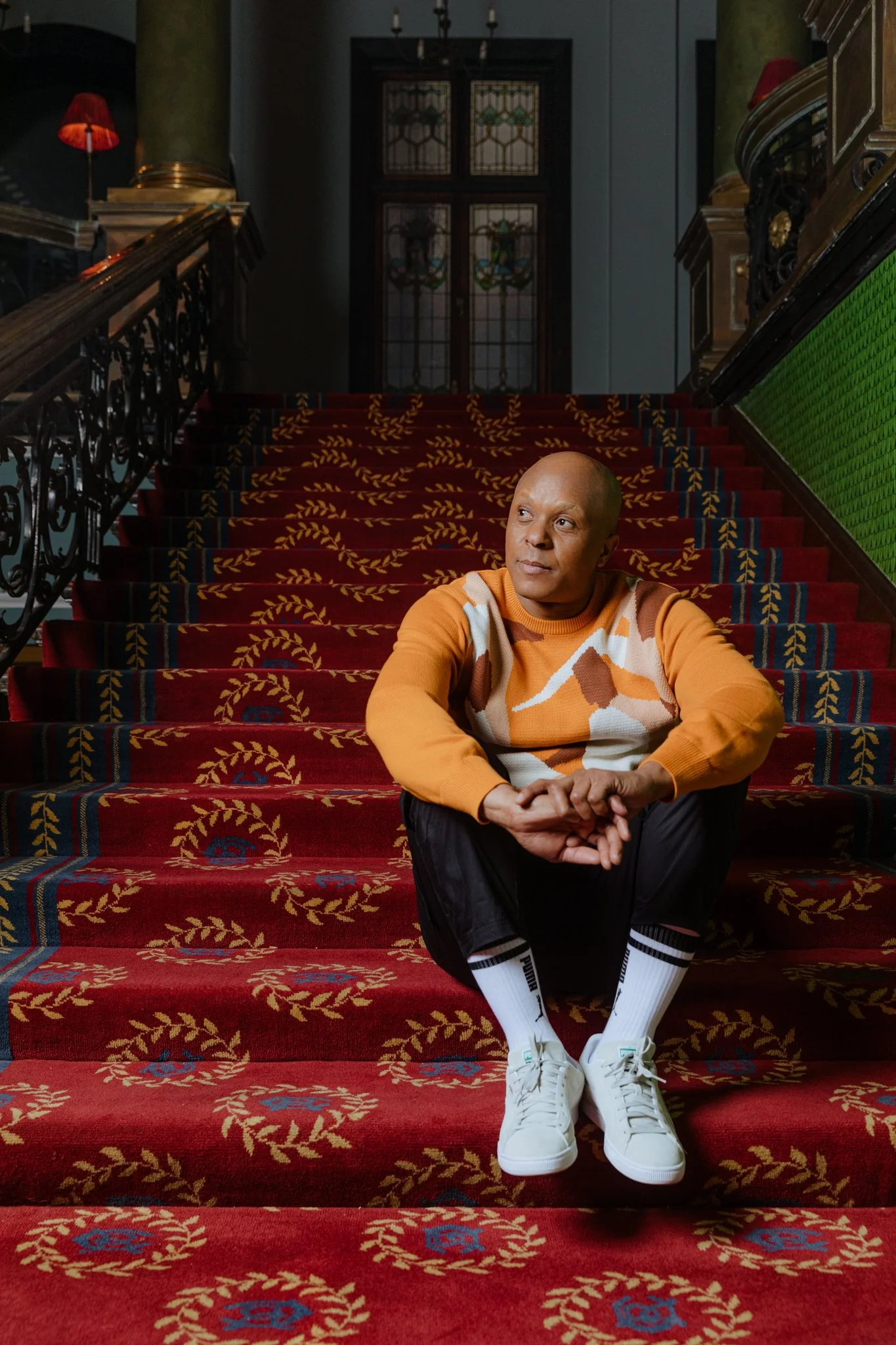 A man sitting on a grand red-carpeted staircase with ornate gold and blue patterns, in a historic building with stained glass windows behind him, wearing an orange sweater, white sneakers, and white socks with black stripes.